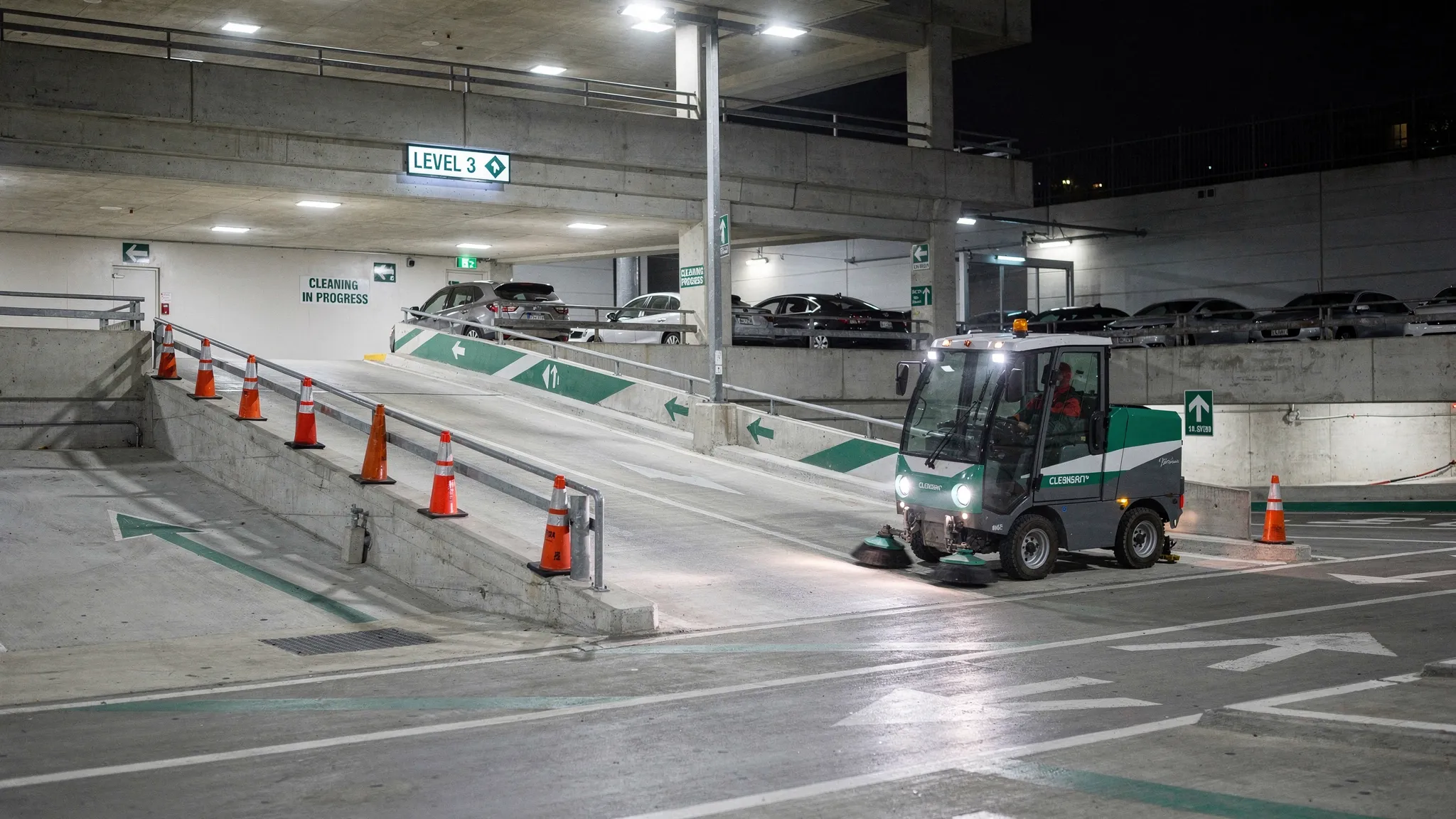 A multi-level concrete parking garage at night with a professional sweeper vehicle cleaning a ramp while safety cones mark a closed lane, overhead lights illuminate dust-free drive lanes, and the scene shows clear signage and a controlled work zone.