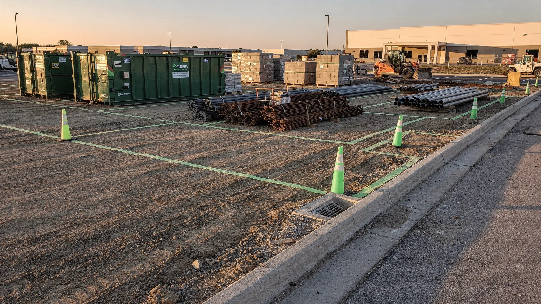A clean commercial construction pad at end of shift with marked travel lanes, dumpsters not overflowing, materials staged within taped laydown boundaries, and a curb line free of sediment near a storm drain inlet.