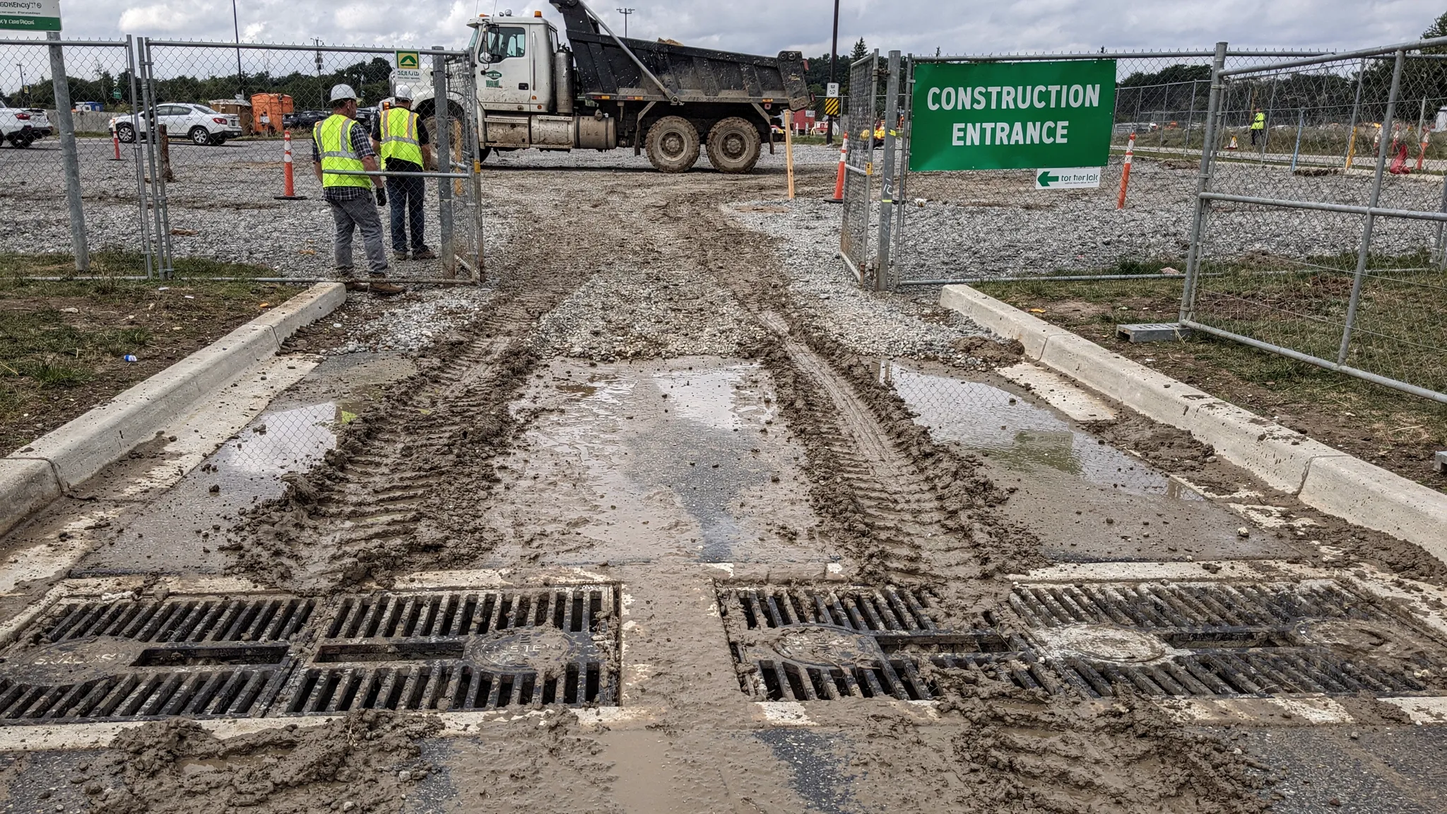 A construction site entrance and haul route with visible mud track-out, curb lines, and nearby storm drain inlets, showing the key areas where sweeping should be timed after rain and heavy trucking.
