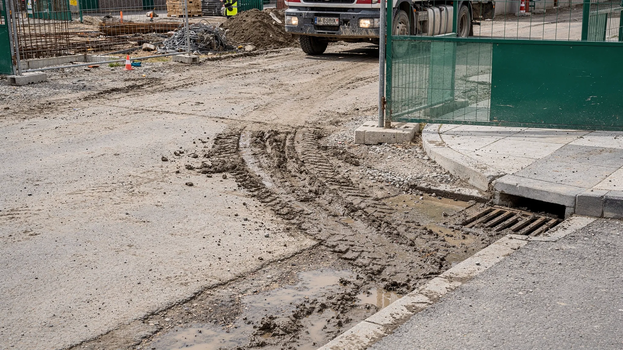 A construction site entrance with visible mud track-out onto a paved public edge and a nearby storm drain inlet, showing why sweeping is needed before inspection. No logos or text on signs.