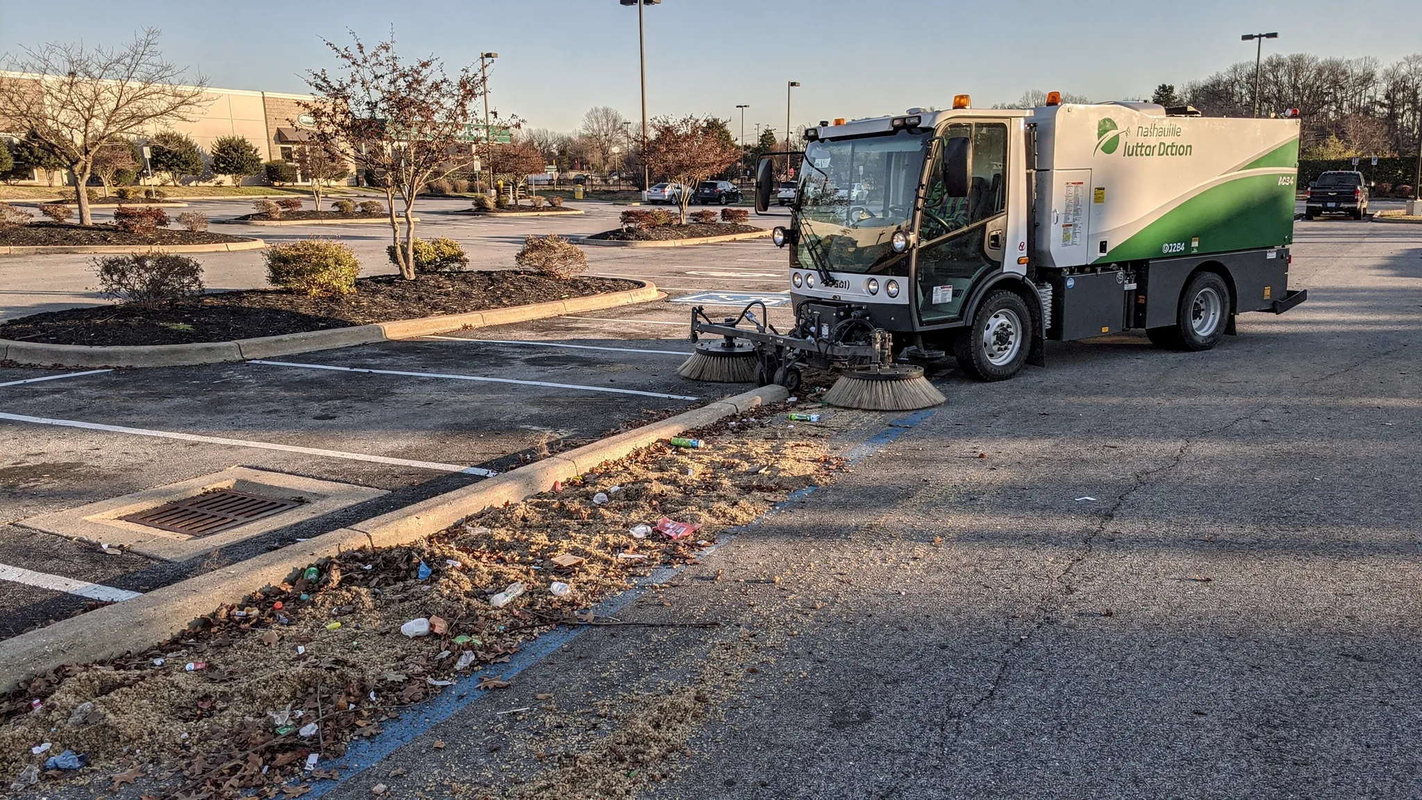 A street sweeper cleaning a commercial parking lot curb line in Nashville, with visible debris piled along the edge before cleaning and a clean curb line behind the sweeper. The scene includes parking islands, storm drain inlet nearby, and clear morning light.