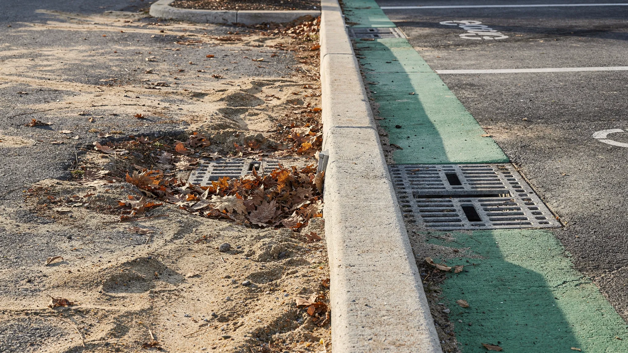 A curb line and storm drain inlet at the edge of a parking lot with visible sediment and leaves on one side and a clean, detailed curb-and-gutter result on the other side, illustrating measurable cleaning outcomes.