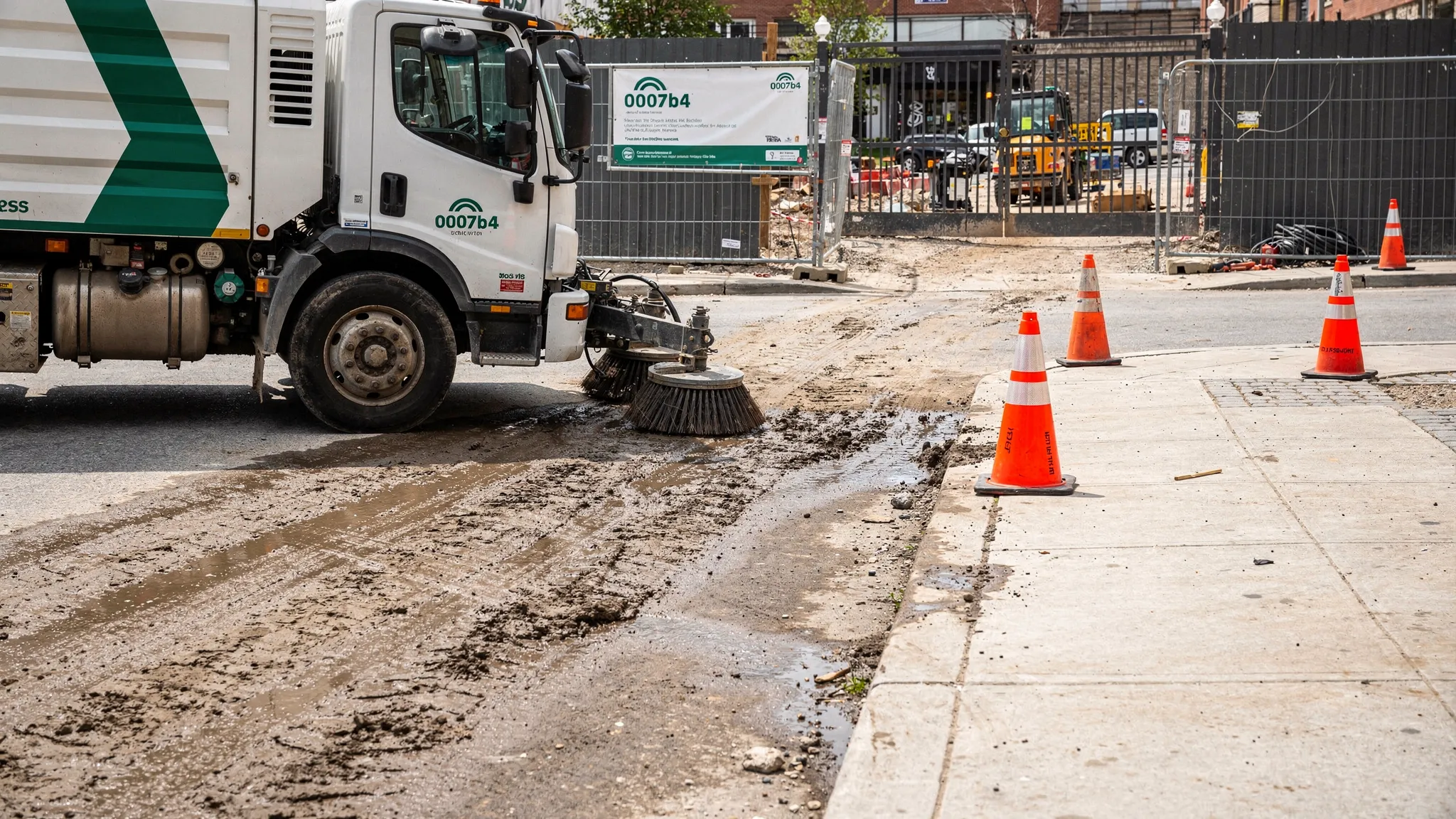 A professional street sweeper cleaning along a curb line near a construction site entrance with visible mud track-out, while safety cones mark the work zone on the roadway.
