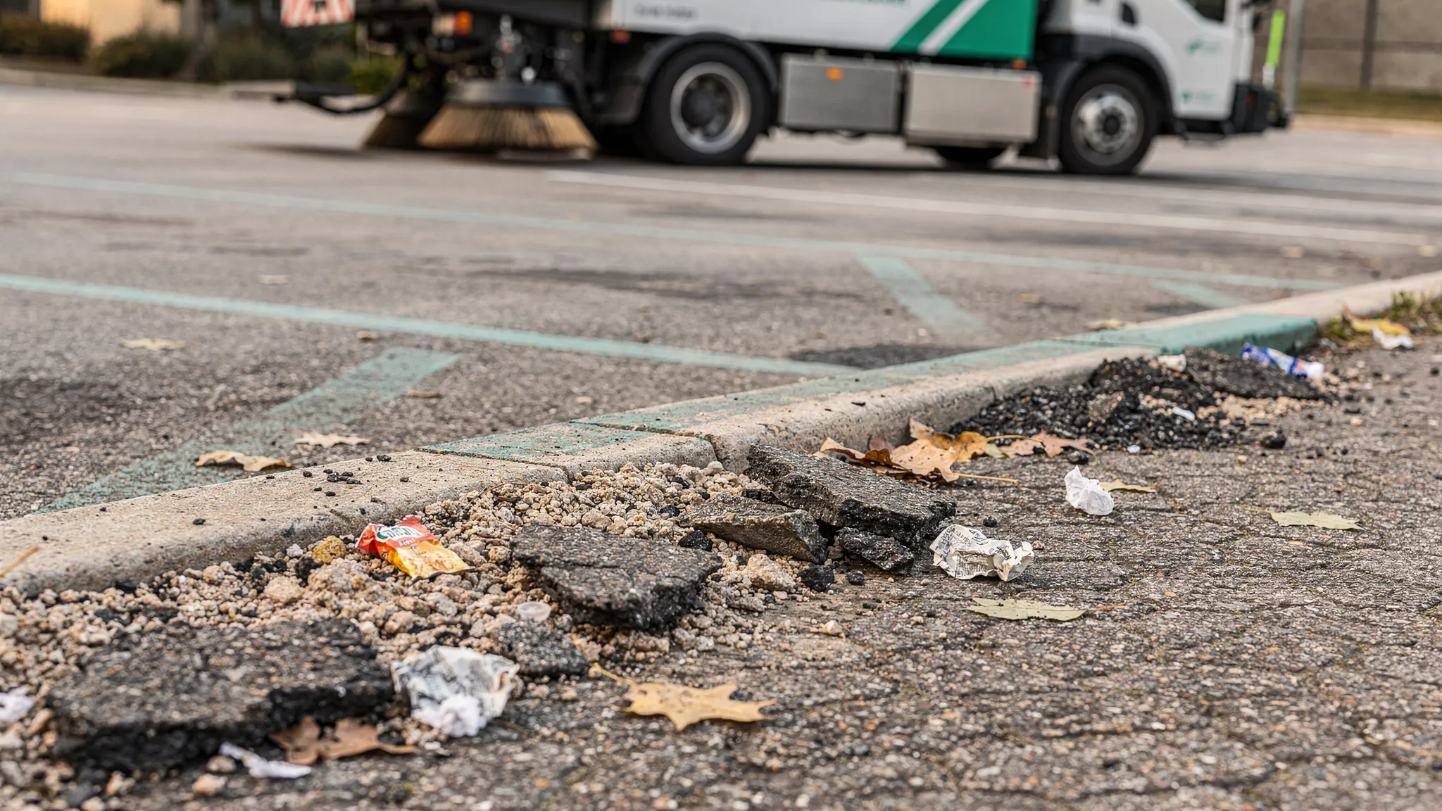 A commercial parking lot edge with visible piles of loose aggregate, small chunks of asphalt, and scattered litter near a curb line, with a street sweeper positioned in the background ready for cleanup.
