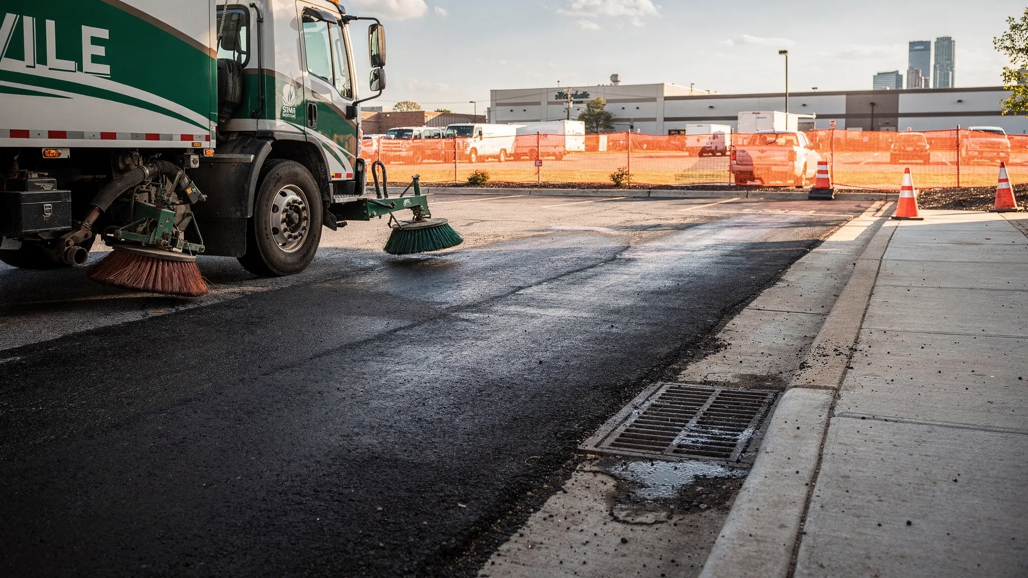 A street sweeping truck cleaning a curb line beside a Nashville commercial parking lot, with a storm drain inlet, construction fencing, and visibly cleaner pavement behind the sweeper.