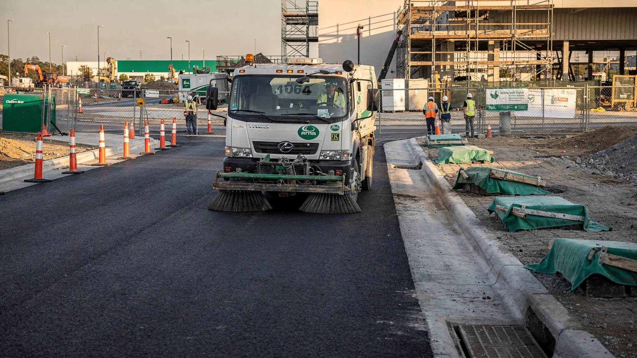 A commercial construction site entrance with a sweeper truck cleaning a paved access lane, cones guiding traffic, cleared curb lines, and protected storm drain inlets near the roadway.