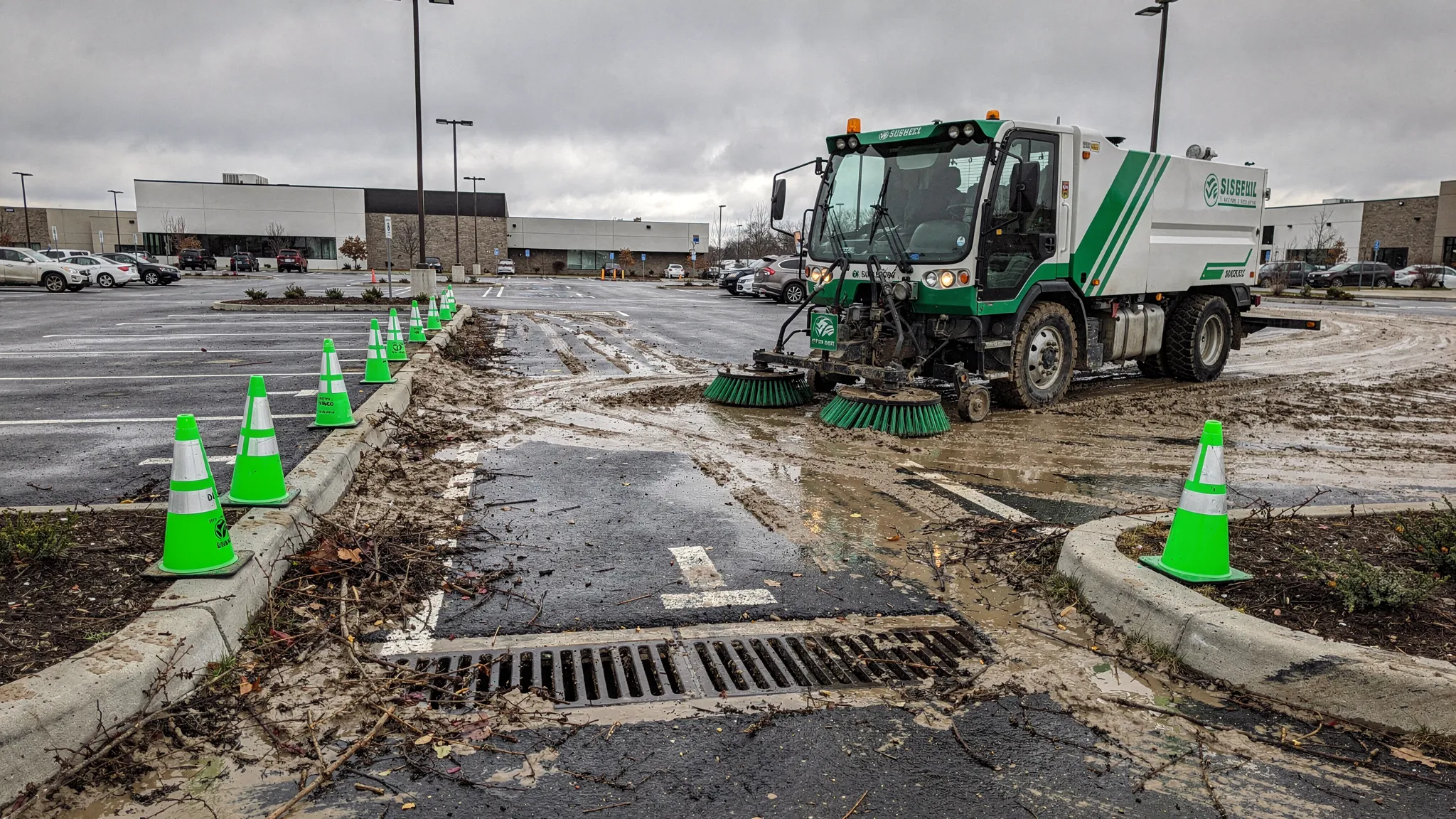 A street sweeper cleaning a storm-affected commercial parking lot with cones marking a muddy drive lane, curb lines being cleared, and storm drain areas kept free of debris.