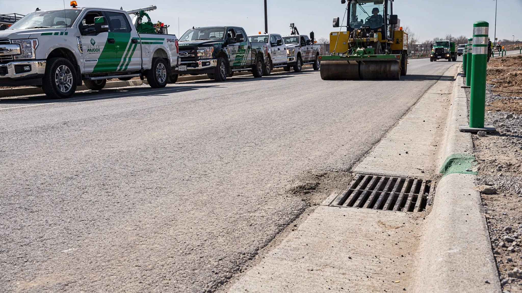 A construction site access road being swept near a curb line, with a clean storm drain inlet nearby, parked work trucks in the background, and dust controlled on the paved surface.