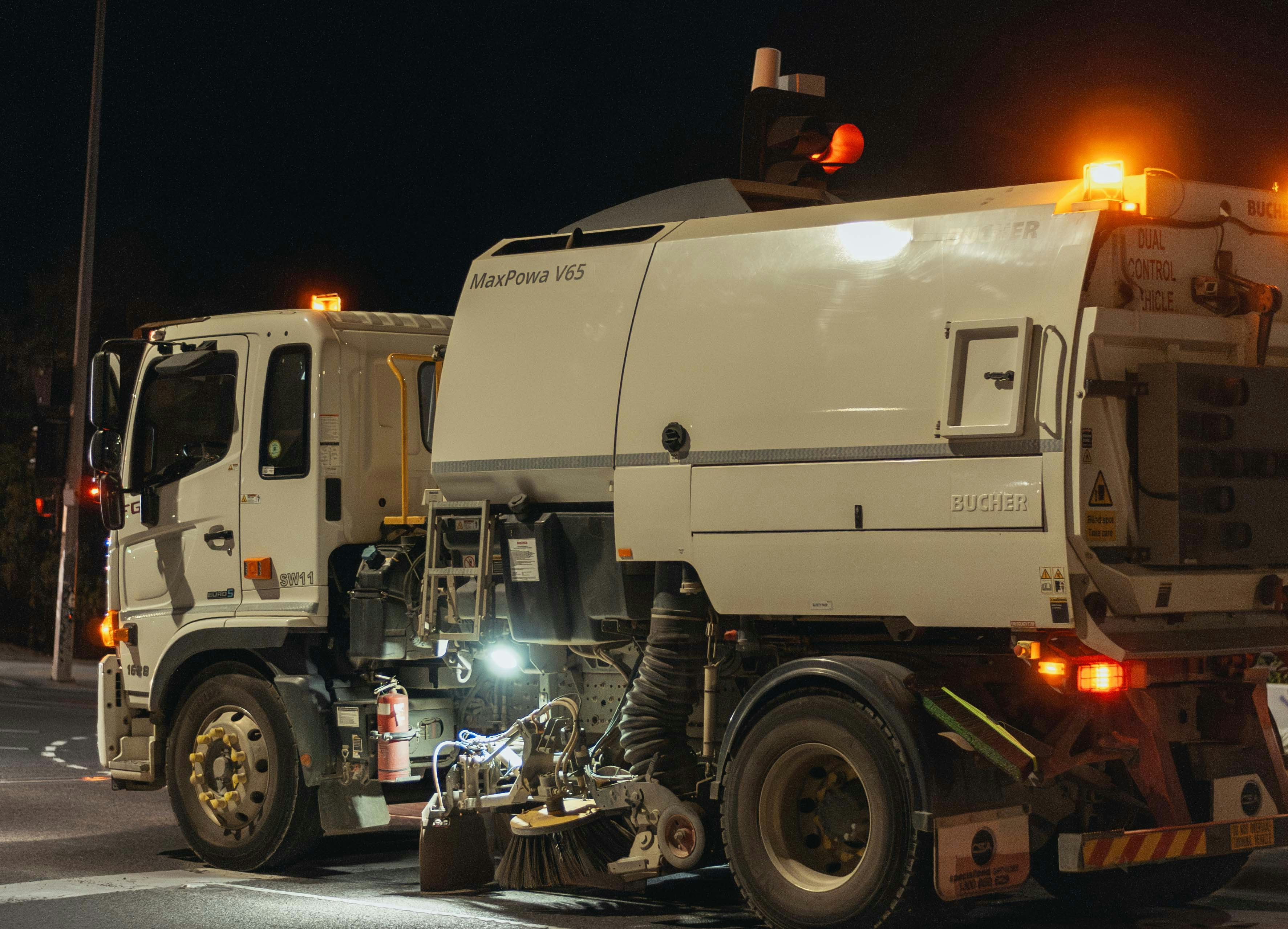 Street sweeper truck operating at night with its lights on, cleaning a city street