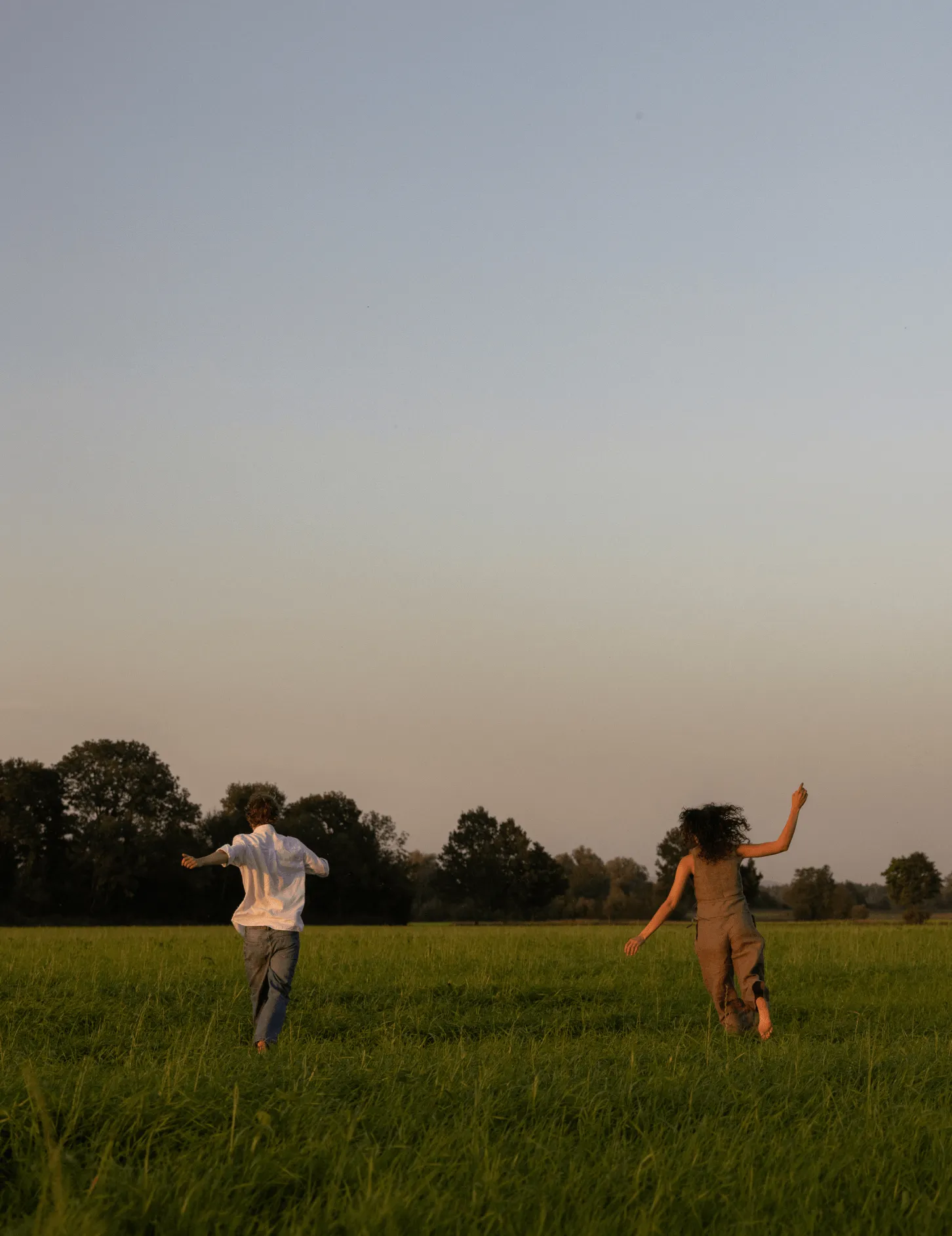 Two people running barefoot through a green grassy field at sunset with trees in the background.