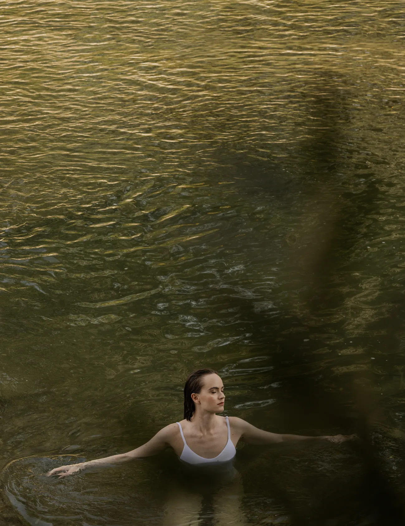 Woman in white swimsuit standing in calm water with eyes closed and arms extended.