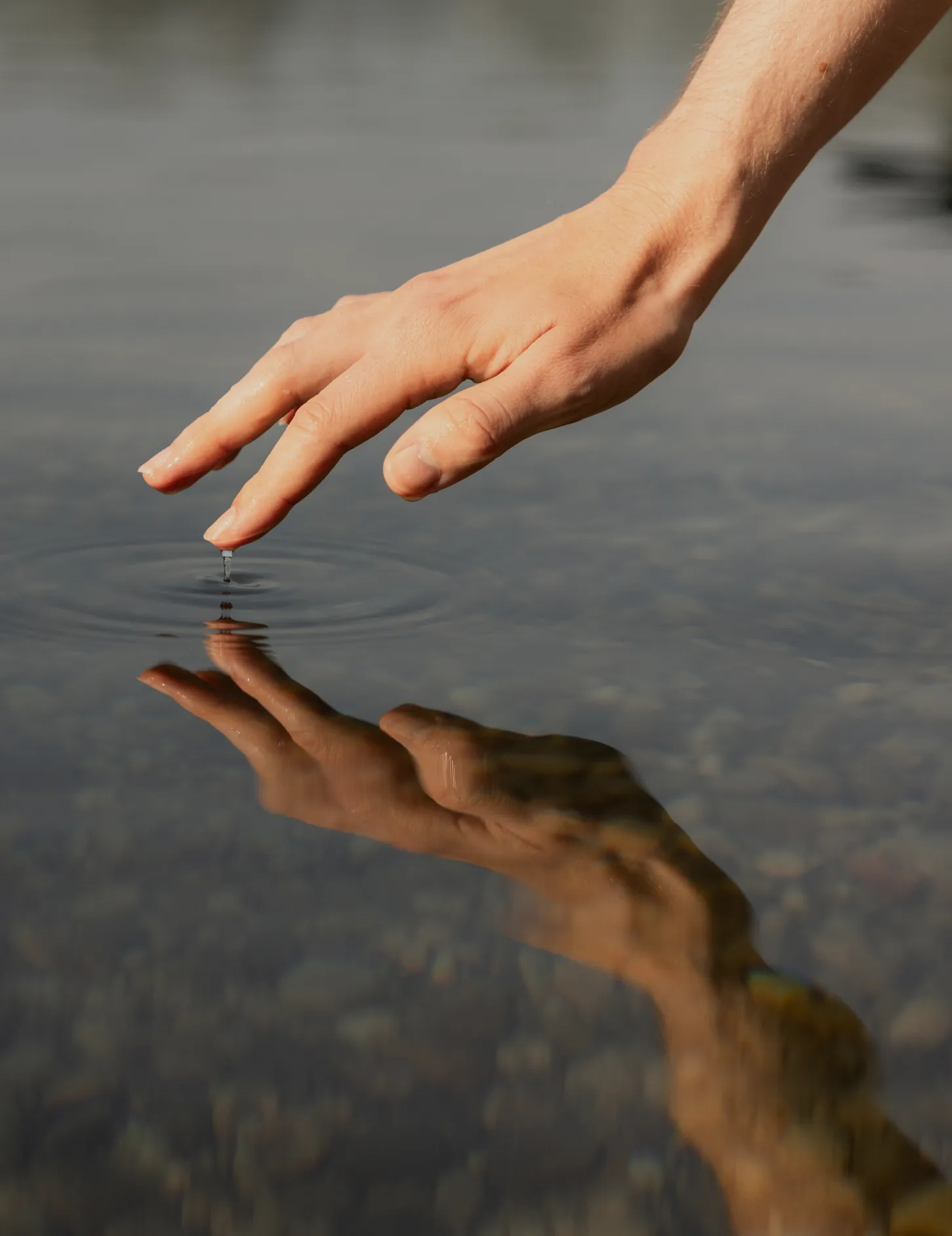 Close-up of a hand with fingers gently touching a calm water surface, creating a small ripple and a visible reflection.
