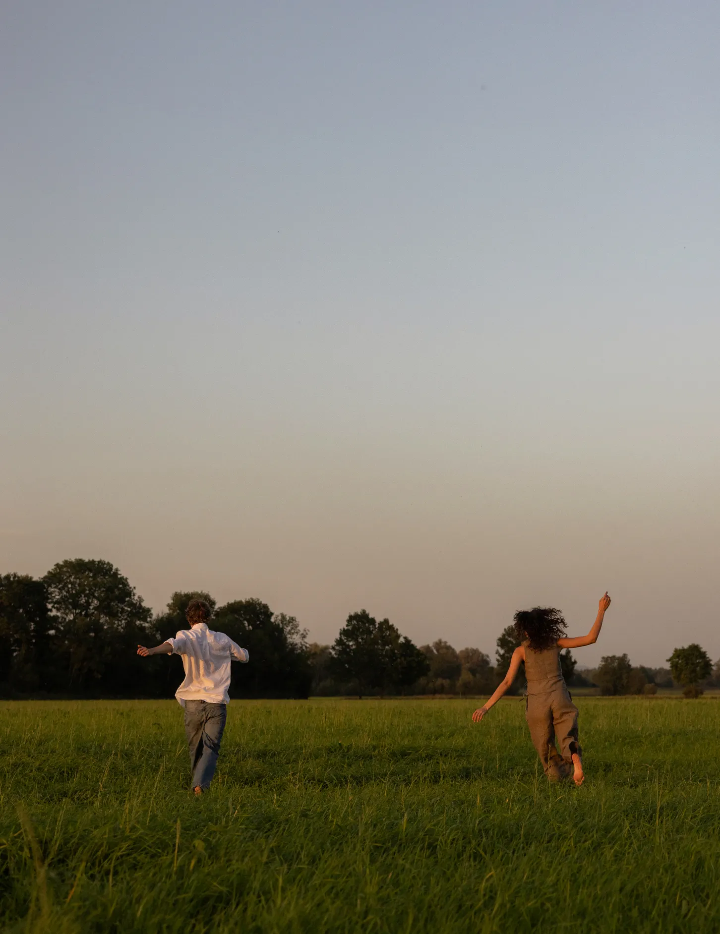 Two people running barefoot through a green field with trees in the distance under a clear sky.