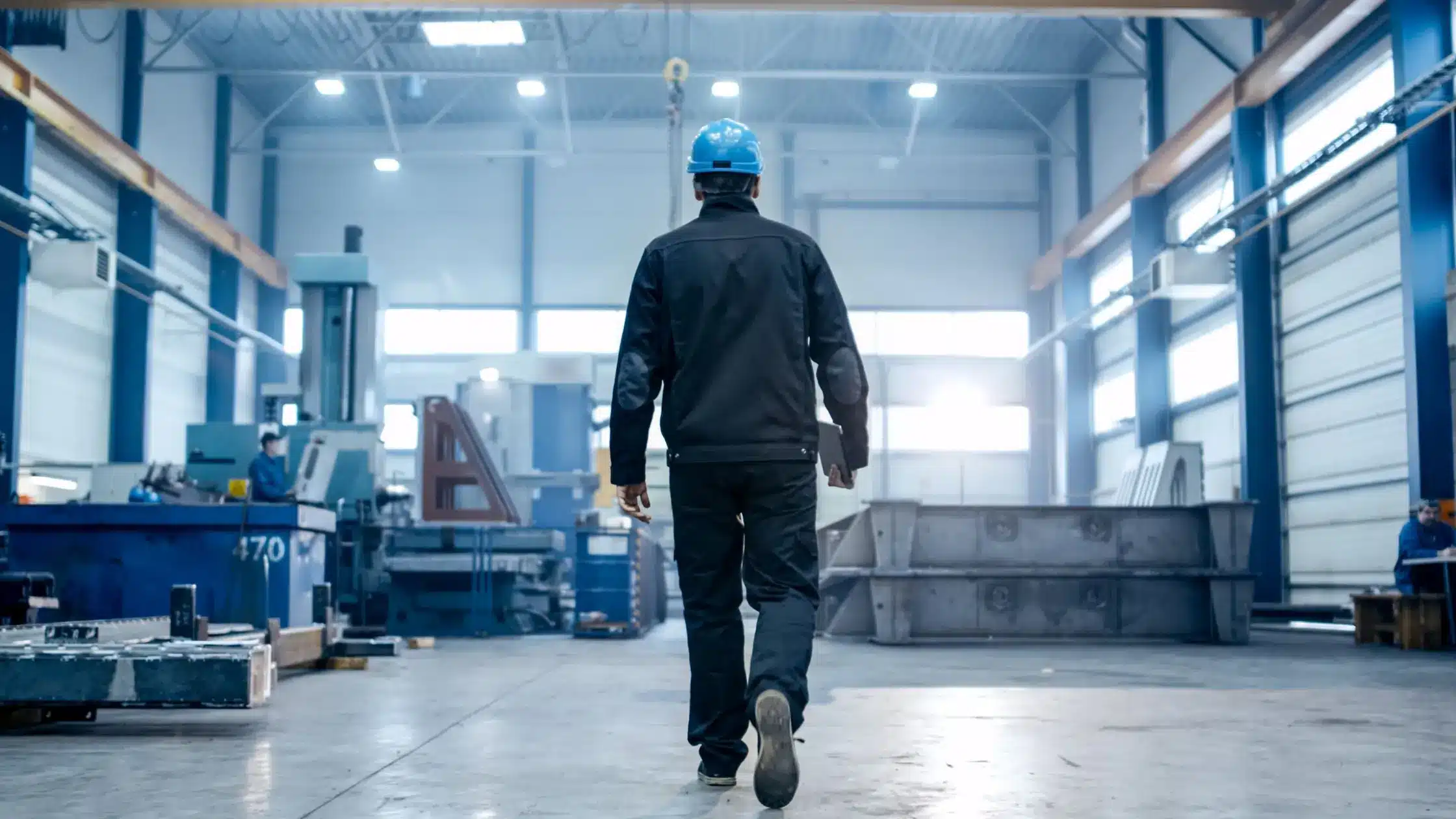 Worker wearing a blue safety helmet walking inside a spacious industrial warehouse with machinery and equipment.