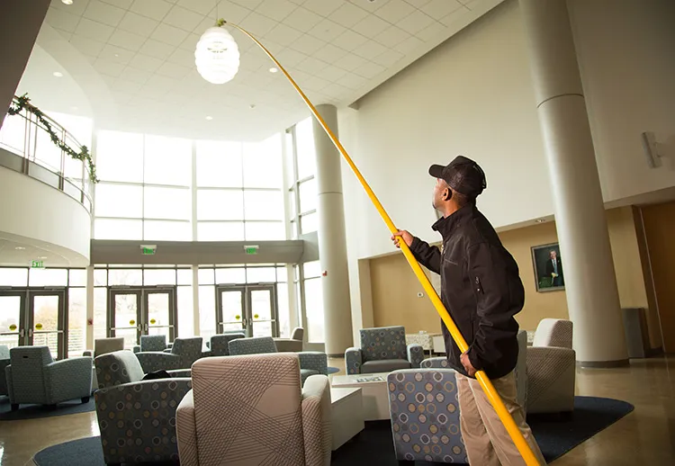 Man using a long yellow pole to clean or adjust a ceiling light in a large, modern lobby with tall windows and multiple chairs.