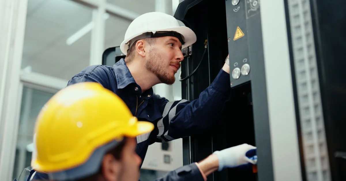 Two maintenance workers wearing hard hats and coveralls inspecting electrical equipment inside a control panel.