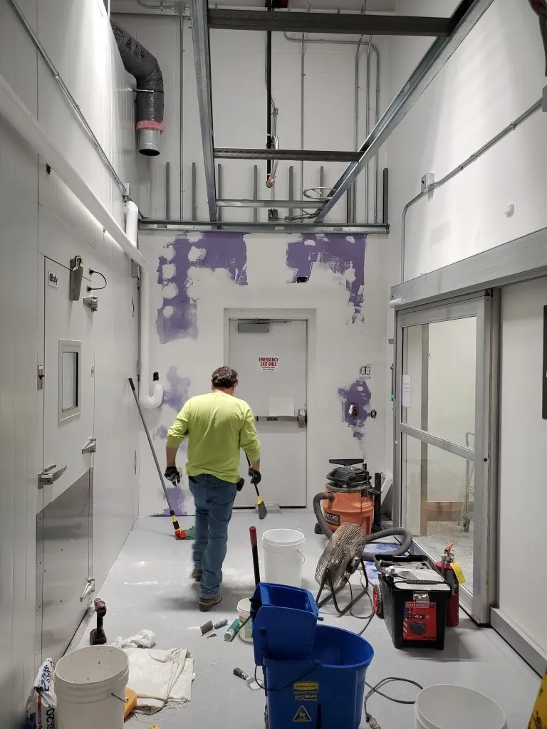 Worker in a neon green shirt working in a room with white walls partially painted purple, surrounded by cleaning and construction equipment.