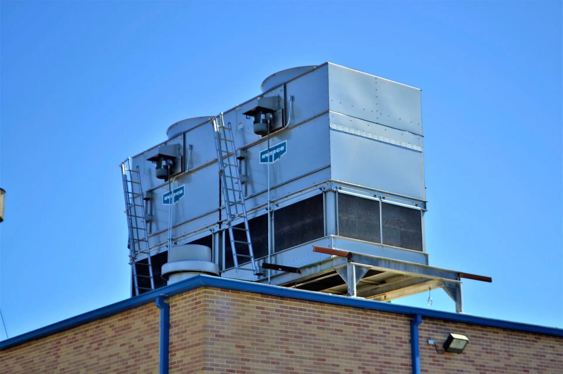 Two large silver HVAC units with ladders mounted on the roof of a brick building under clear blue sky.
