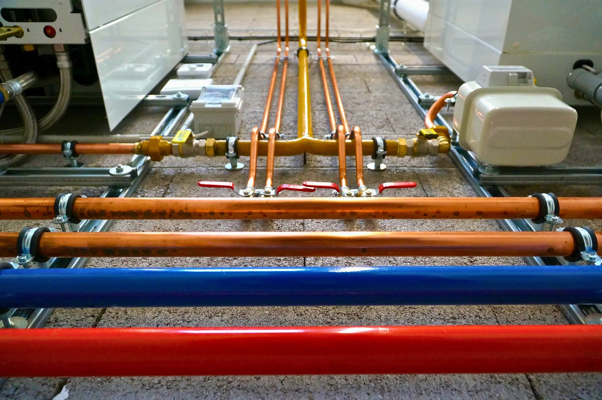 Close-up view of various colored pipes, including red, blue, and copper, organized on a floor with metal clamps and valves.