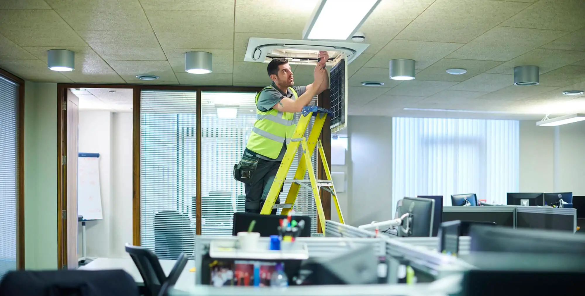 Maintenance technician standing on a yellow ladder repairing a ceiling air conditioning unit in a modern office space with desks and computers.