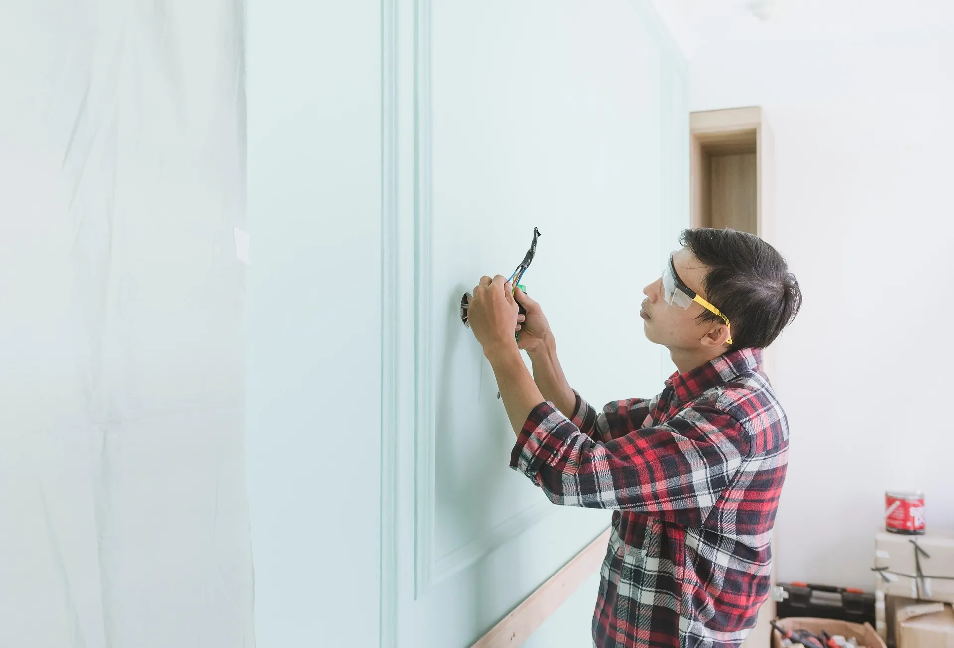 Man wearing protective glasses working on electrical wiring inside a light blue paneled wall.