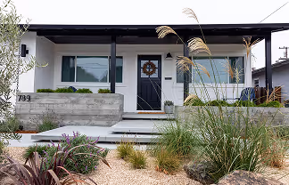 Modern white house front with black door decorated with a wreath, large windows, concrete planters, and landscaped garden with grasses and rocks.