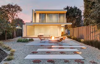 Modern two-story house with large windows, outdoor seating, and a fire pit on a gravel patio at sunset.