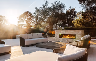 Outdoor patio with modern sectional sofa and a gas fireplace at sunset surrounded by trees.