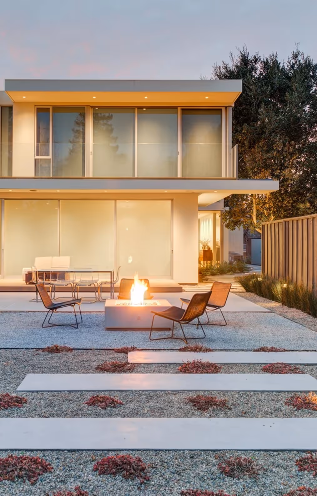 Modern two-story house with large glass windows, patio seating, and a lit fire pit in the backyard at dusk.