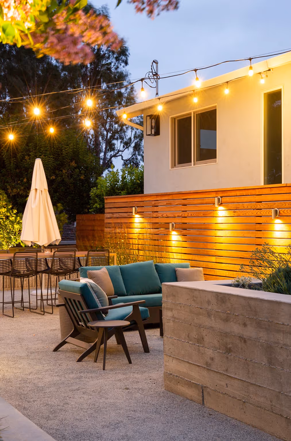 Cozy outdoor patio with teal cushioned chairs, string lights, wooden fence, and dining table with umbrella at dusk.