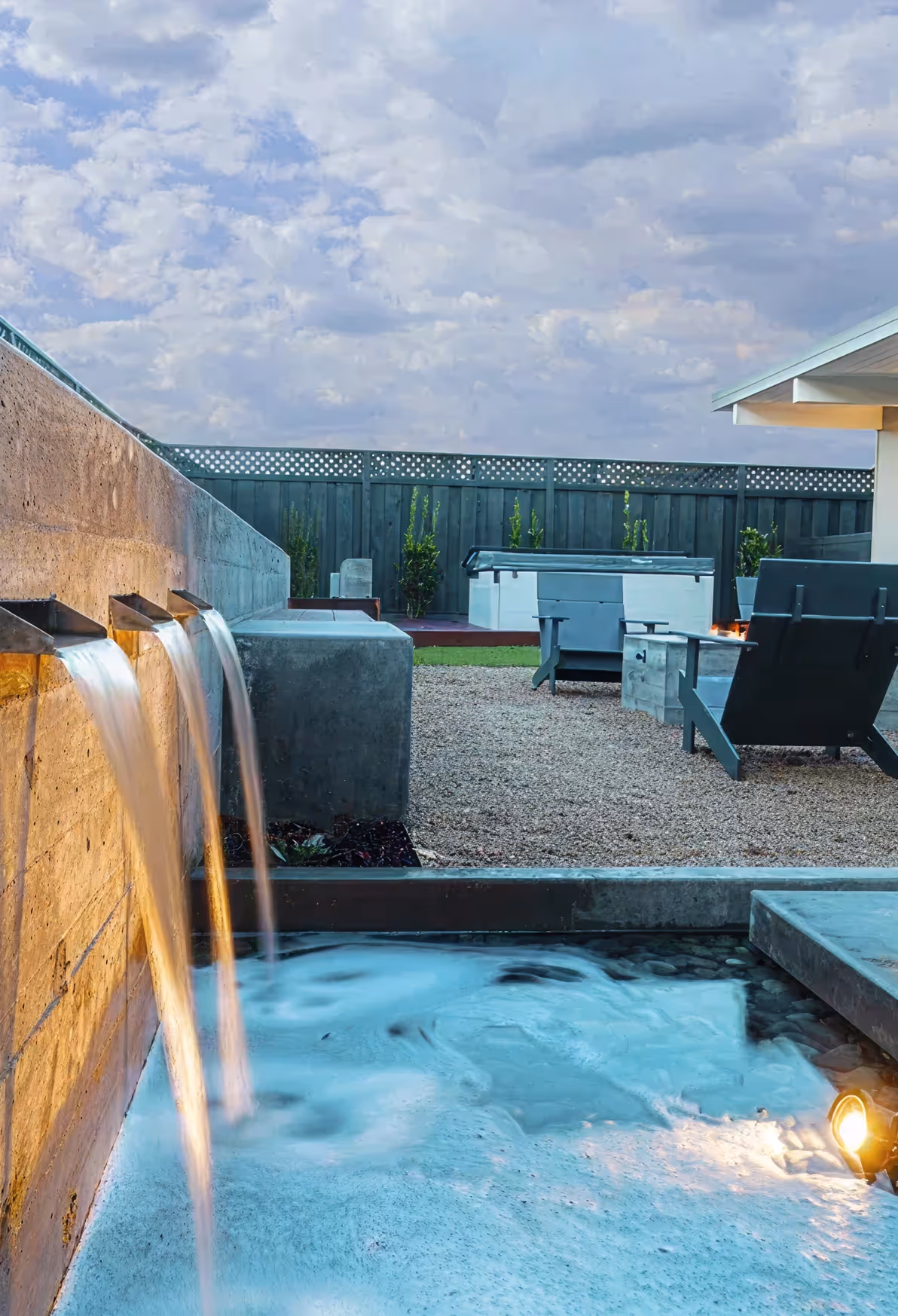 Modern backyard featuring a concrete wall with three spouts releasing water into a shallow pool, surrounded by gravel, Adirondack chairs, a fire pit, and a hot tub against a wooden fence under a cloudy sky.