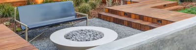 Outdoor seating area with a metal bench, circular fire pit filled with stones, and wooden decking surrounded by gravel and plants.