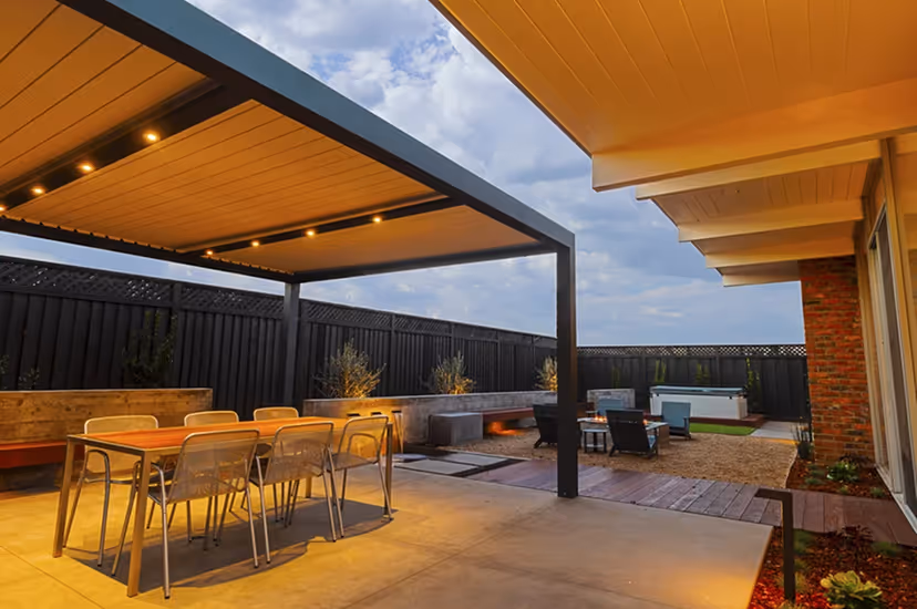 Modern outdoor patio with a covered dining area featuring a wooden table and metal chairs, adjacent to a fire pit and seating area under a partly cloudy sky.