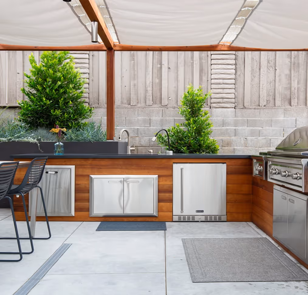 Modern outdoor kitchen with wooden cabinets, stainless steel appliances, bar stools, and green plants under a white canopy.