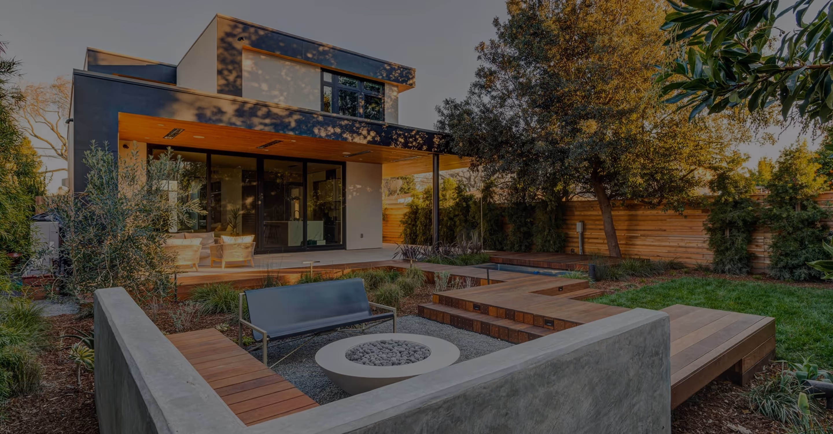 Modern backyard with a fire pit surrounded by wooden benches and a metal bench, next to a two-story house with large windows and a covered patio.