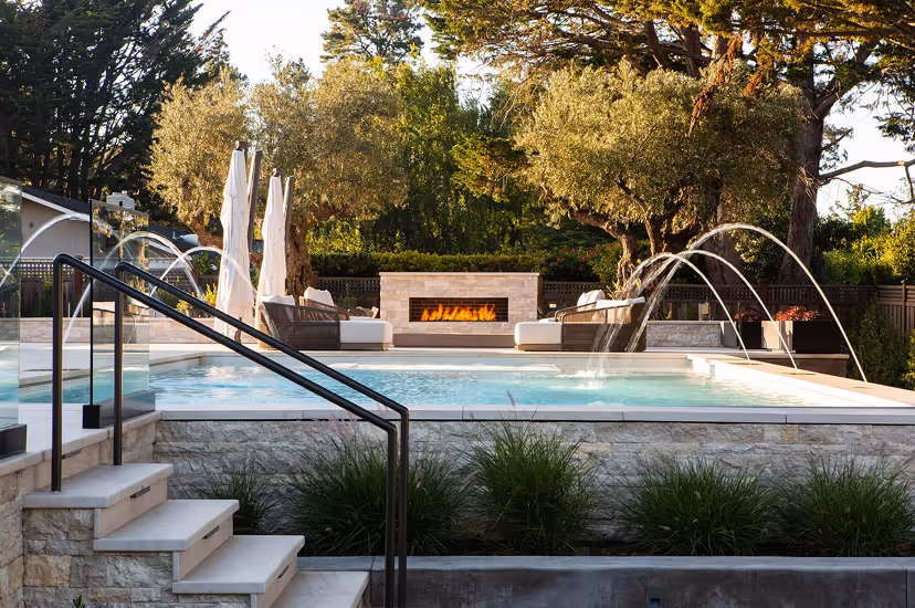 Modern outdoor pool area with a stone fireplace, water fountains arcing into the pool, cushioned seating, and surrounding trees.