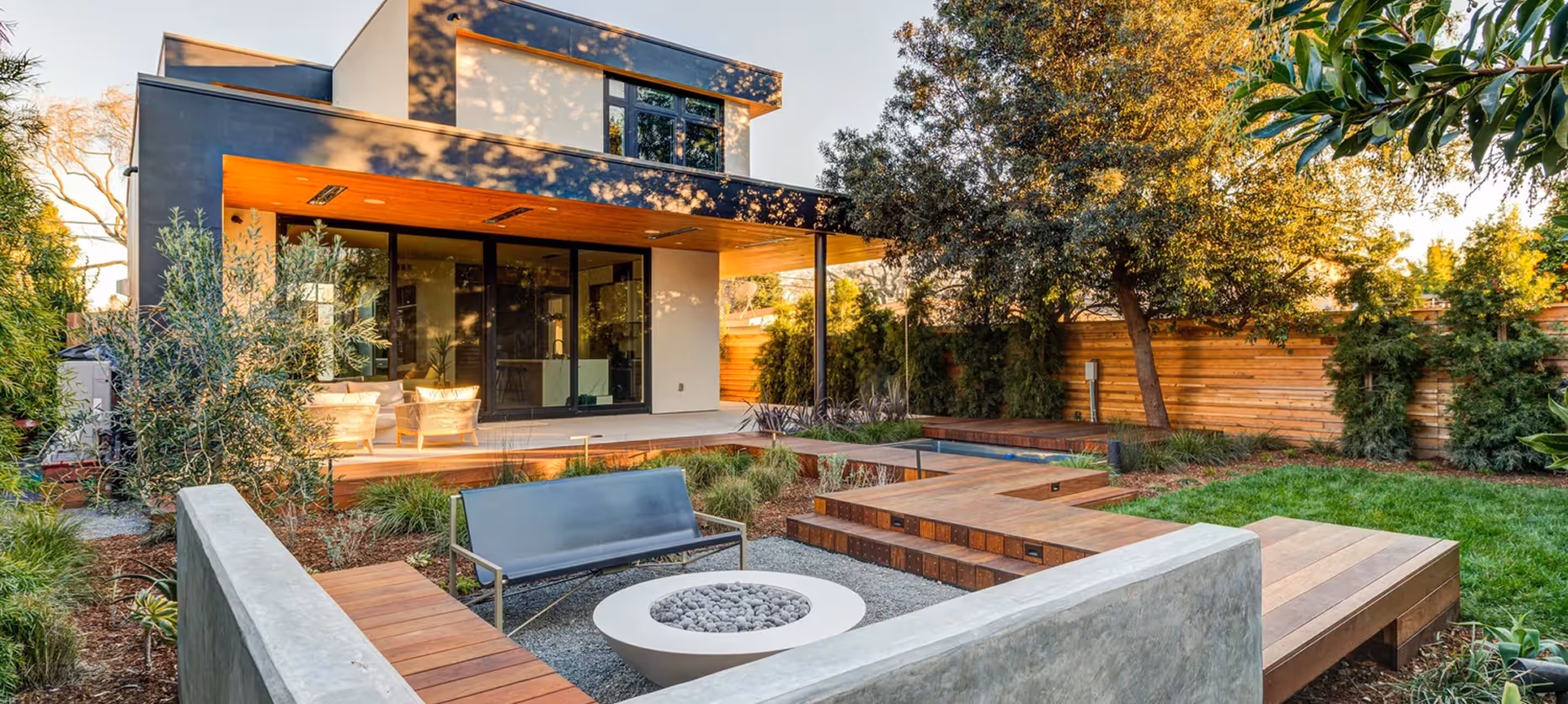 Modern backyard with a wooden deck, outdoor seating, a circular fire pit with stones, concrete and wood benches, and green lawn surrounded by trees and a wooden fence.