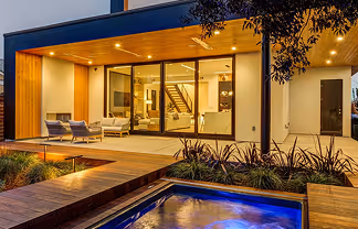 Modern house exterior at dusk with illuminated porch, glass windows, and a lit water feature in the foreground.