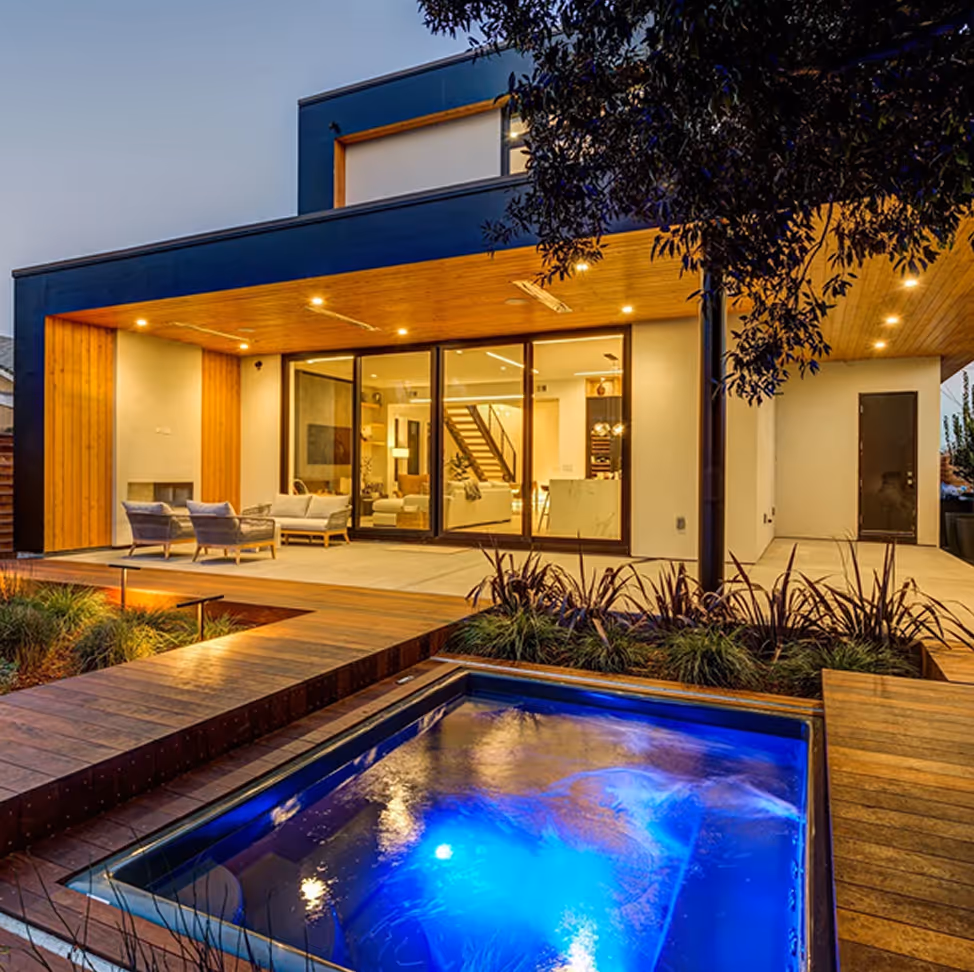 Modern house with wooden accents, outdoor seating area, and illuminated blue water feature at dusk.