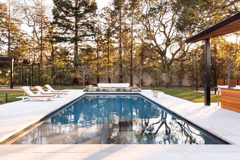 Modern rectangular outdoor swimming pool surrounded by white stone patio, lounge chairs, and trees.