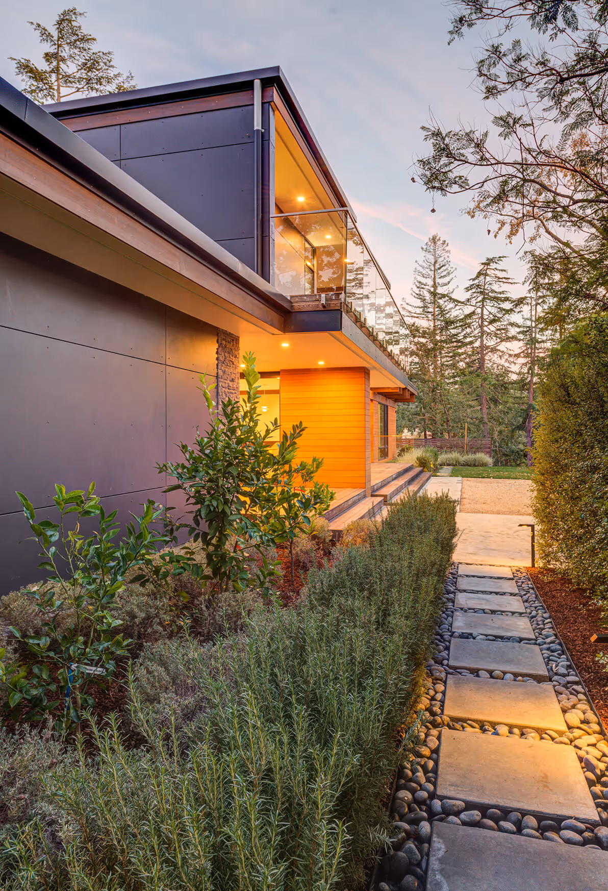 Modern house exterior with black and wooden panels, glass balcony, and a stone path lined with shrubs and trees at sunset.