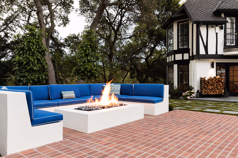 Outdoor patio with a white L-shaped sofa featuring blue cushions surrounding a rectangular fire pit, set on a brick floor with trees and a Tudor-style house in the background.