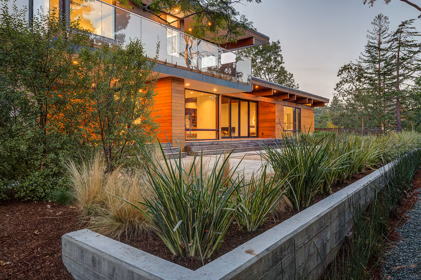 Modern two-story house with wooden paneling, large glass windows, and a balcony overlooking a garden with ornamental grasses and shrubs.