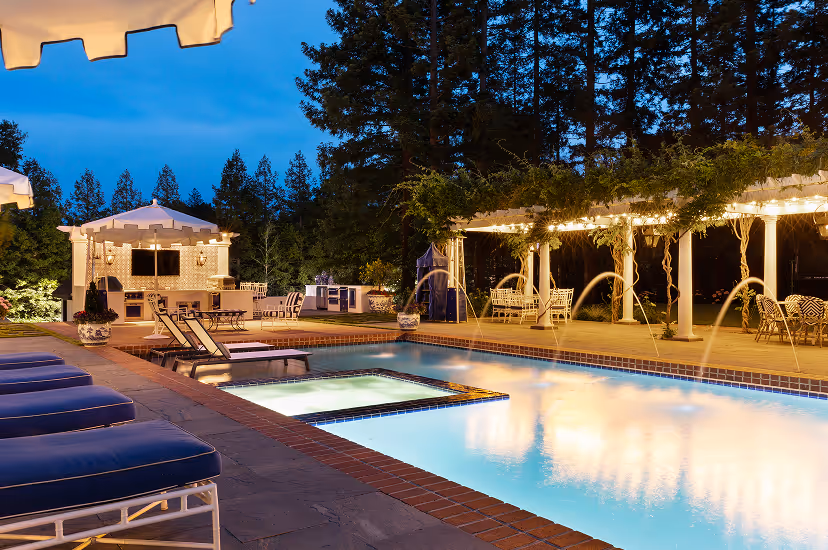 Outdoor swimming pool area at dusk with lounge chairs, illuminated water fountains, and a covered seating area with string lights.
