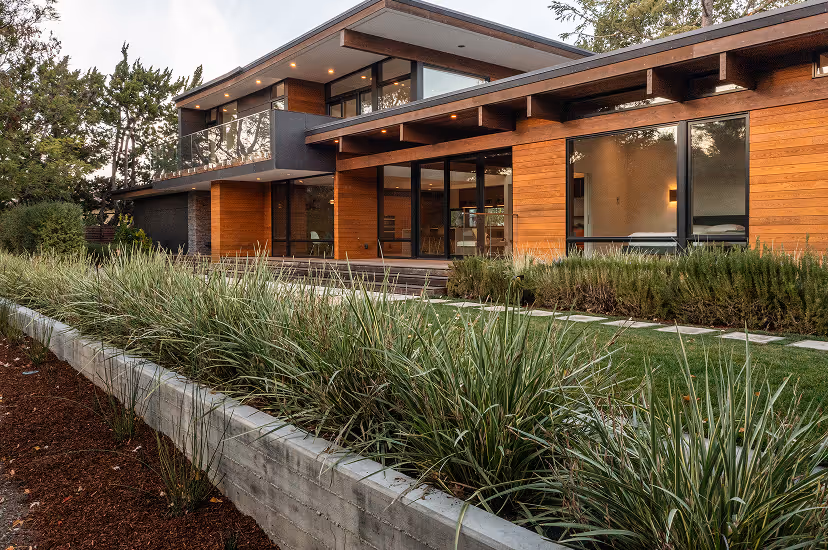Modern two-story house with wooden panel exterior, large glass windows, and landscaped garden with green shrubs and grass.
