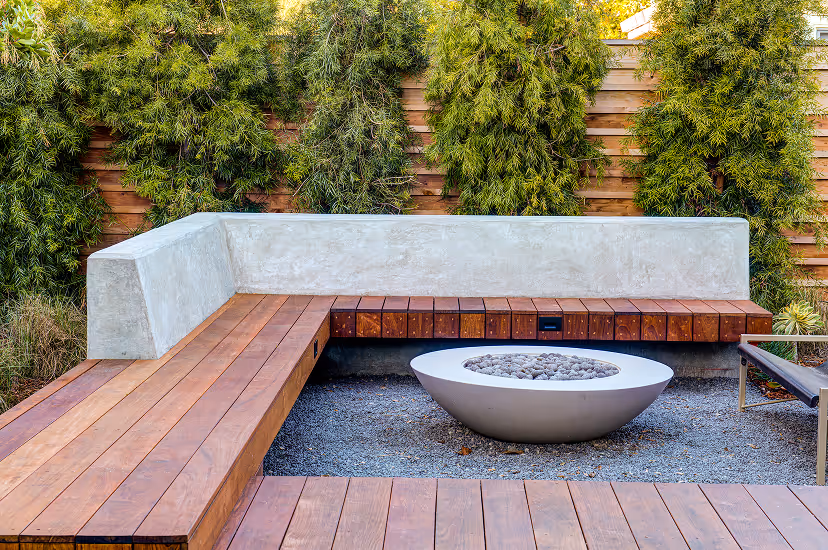 Modern outdoor seating area with wooden benches, concrete backrest, round fire pit, and green plants along a wooden fence.