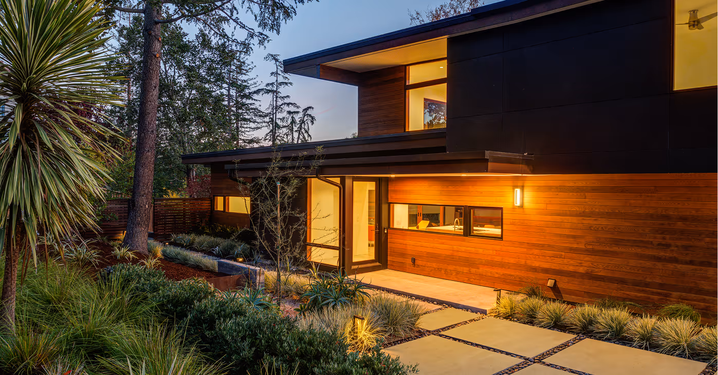Modern two-story house exterior at dusk with wood and black panel siding, illuminated entrance, and landscaped garden with various plants.