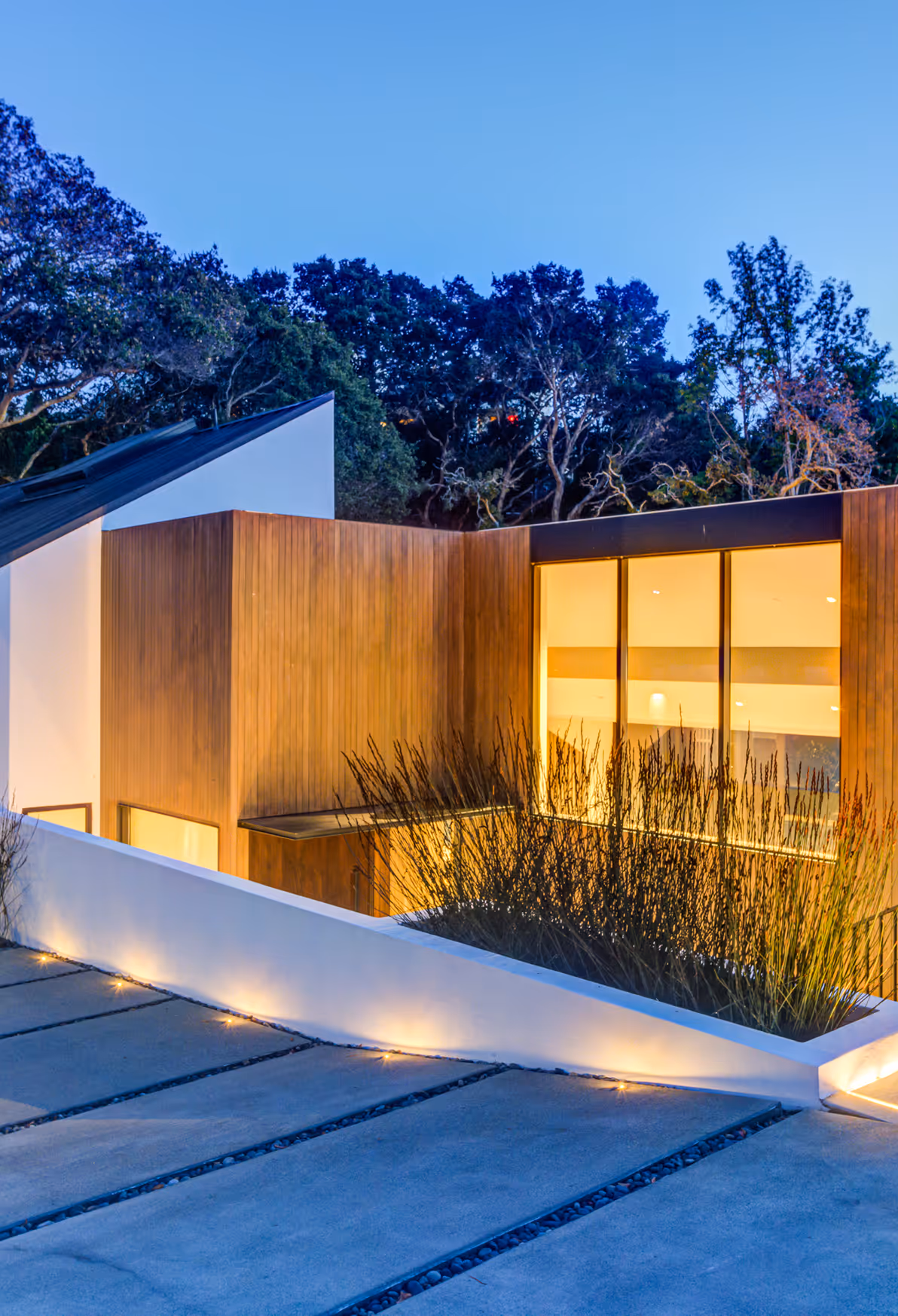 Modern house exterior at dusk with wood paneling, large illuminated windows, and a concrete driveway with embedded lights.