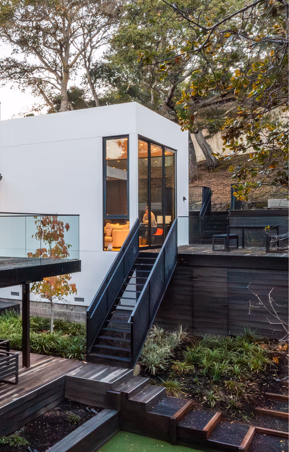 Modern white house with large black-framed windows, outdoor stairs, and surrounding greenery in a wooded area.