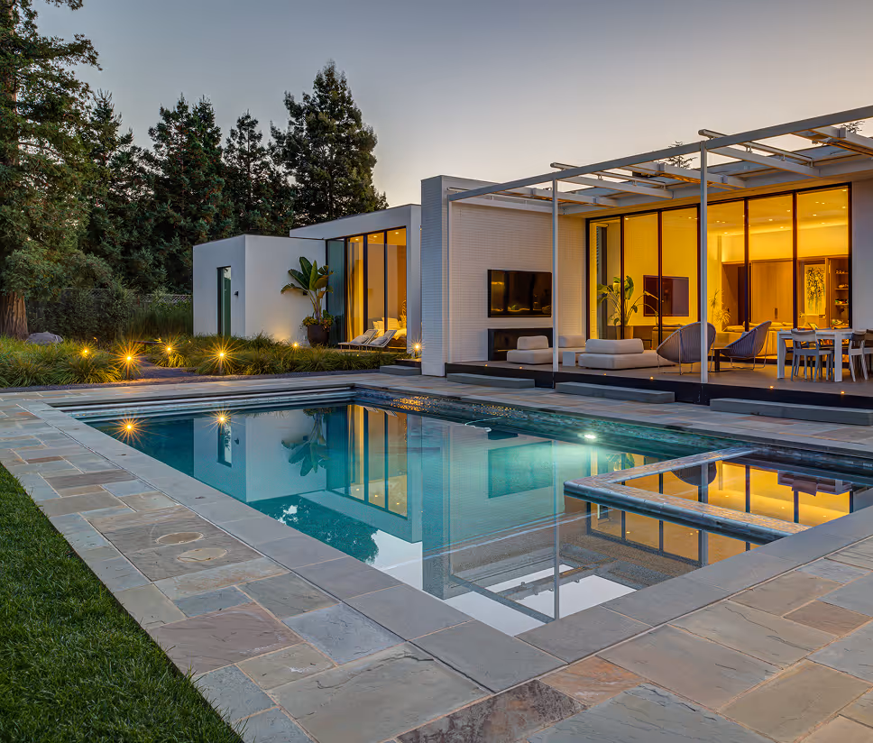 Modern house with large glass windows and sliding doors opening to a poolside patio and swimming pool at dusk.